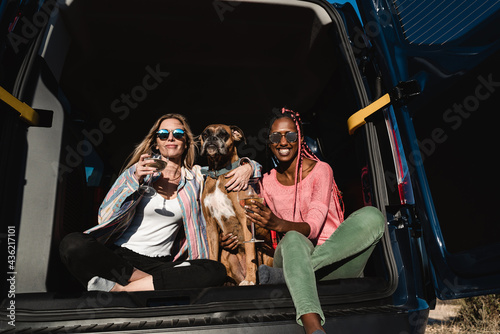 Hipster friends with dog celebrating with wine together in camper van during summer travel vacation - Main focus on african girl face