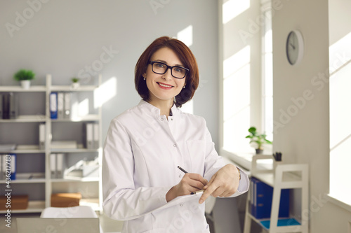 Medical staff. Portrait of an experienced female doctor with documents in hands at the clinic office. Woman in a white medical gown and glasses looks at the camera while filling out a medical card.