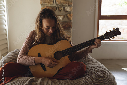 Happy caucasian woman sitting on beanbag playing acoustic guitar in sunny cottage living room