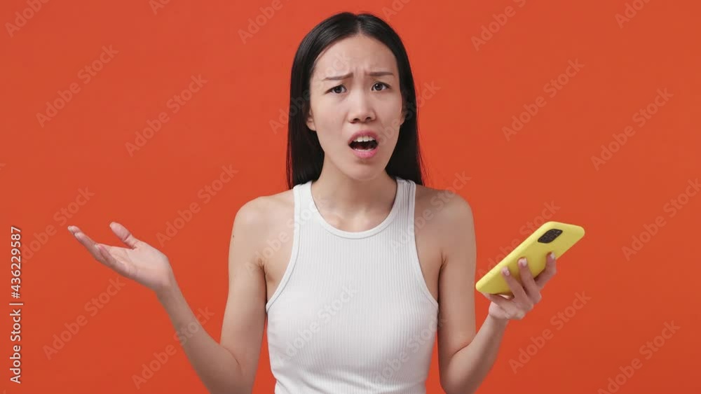 Sad irritated young asian woman 20s years old wears white tank top shirt use mobile cell phone swear worry hears fake news unexpected rumor has some problems isolated on orange color background studio Sad irritated young asian woman 20s years old wears white tank top shirt use mobile cell phone swear worry hears fake news unexpected rumor has some problems isolated on orange color background studio