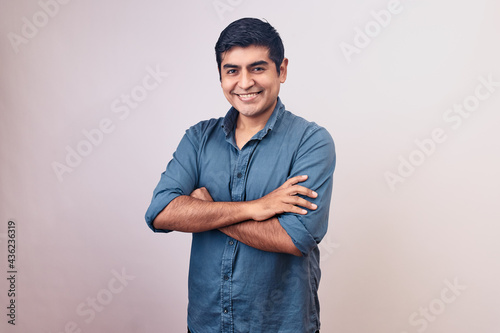 Hombre joven y feliz sonriendo en primer plano cruza brazos. Modelo aislado en fondo blanco con camisa azul