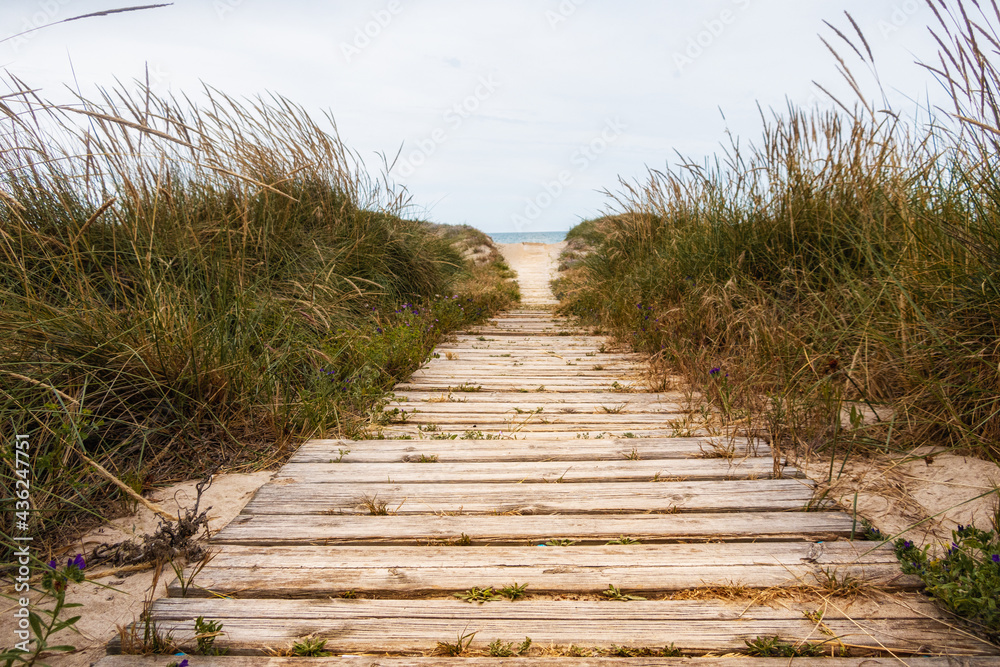 Fototapeta premium Wooden walkway extended on a fine sandy beach with the Mediterranean Sea in the background. In a cloudy sunset.
