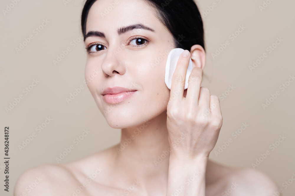 Beautiful Caucasian woman gently cleaning face with makeup remover pad ion beige background