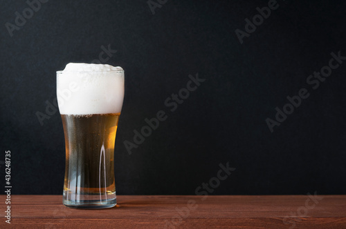 Foamy cold beer in glass beer mug on wooden table in a pub. Dark black wall as background for text, menu, mockup