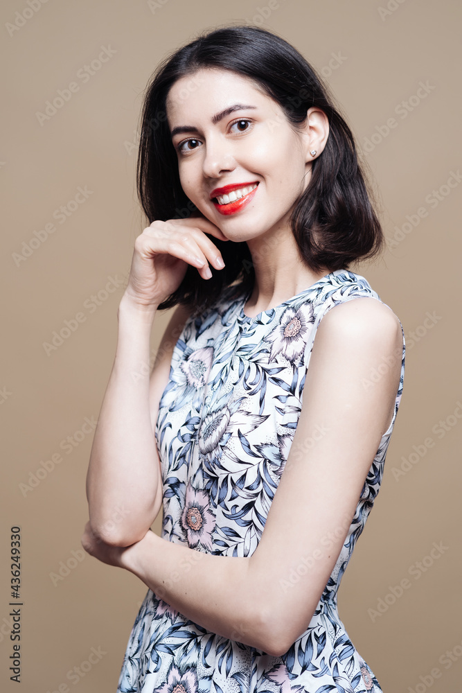 Beautiful young woman in dress with flower print posing cheerfully on beige background Beautiful young woman in dress with flower print posing cheerfully on beige background