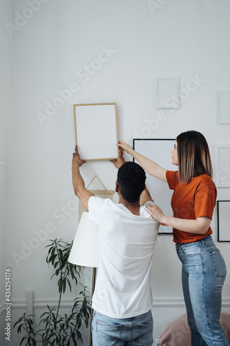Obraz na plátně Excited african young husband and caucasian wife hang frame picture on wall happ