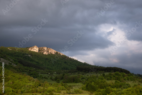 Great, soft light on a pointy, rocky mountain peak and landscape covered by vivid green spring colored forests under a cloudy, moody sky