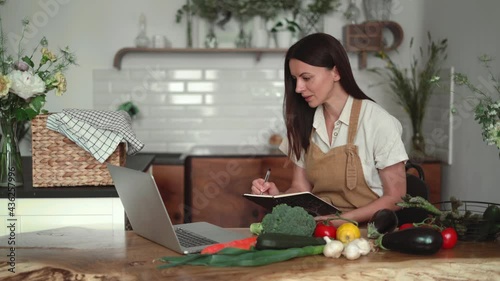 A beautiful young woman prepares from fresh organic vegetables, looking at a laptop screen in the kitchen, writes a recipe in a notebook, dressed in linen clothes. Learning online dietology