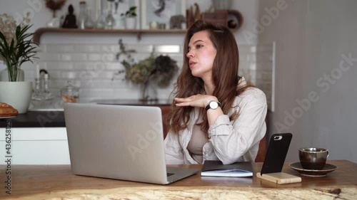 Smiling young blonde woman in beige linen clothes with her laptop computer, drinking coffee, singing while sitting at the table indoors. Homeschooling. Freelancer works from home