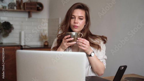 Smiling young blonde woman in beige linen clothes with her laptop computer, drinking coffee, singing while sitting at the table indoors. Homeschooling. Freelancer works from home