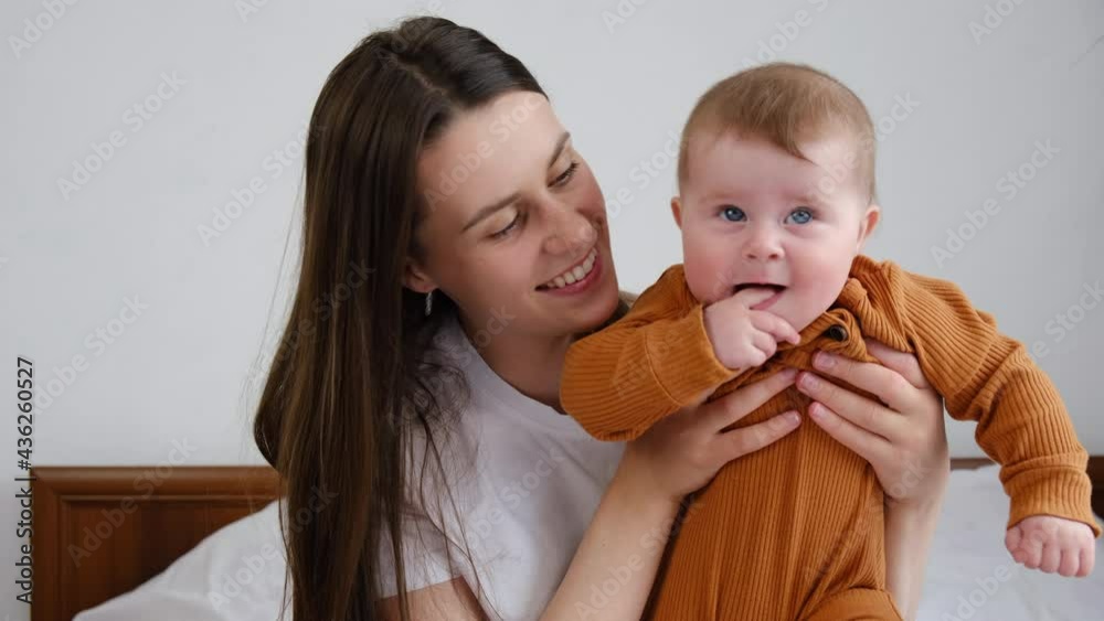 Caring smiling young caucasian mother holds and kissing cute adorable small infant daughter sit on comfy bed at home, looking at camera. Diverse loving happy mom caressing little 7 months baby girl Caring smiling young caucasian mother holds and kissing cute adorable small infant daughter sit on comfy bed at home, looking at camera. Diverse loving happy mom caressing little 7 months baby girl