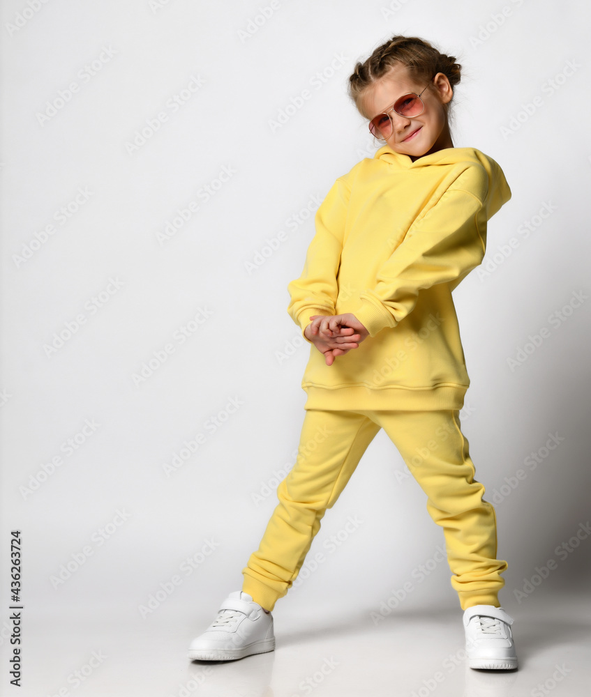 Full length portrait of a little girl dressed in a bright yellow sports suit. Stylish cute child in sunglasses posing on a white background. Concept of active childhood.
