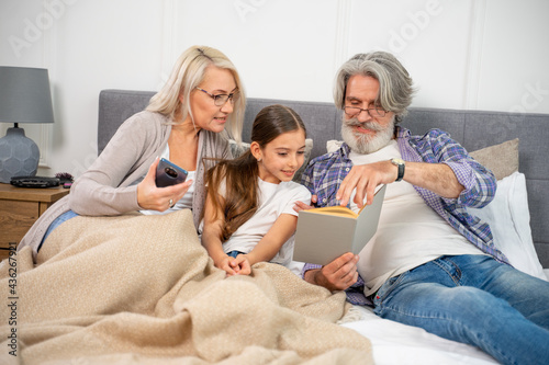 Small little girl granddaughter smiling when reading book with caring senior grandparents while lying on spacious bed enjoying leisure time with grandma and grandpa on weekend at home. Family concept