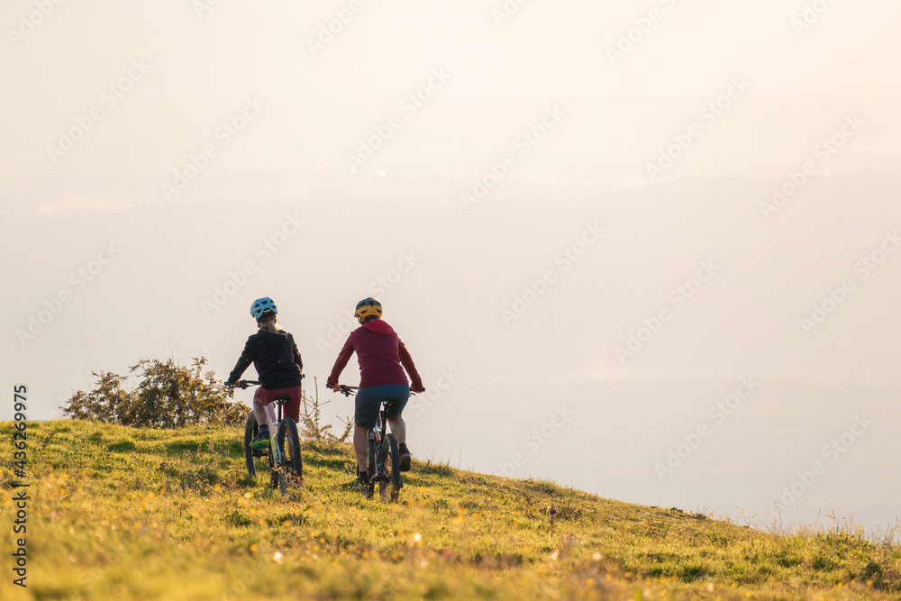 Mother and daughter cycling downhill with mountain bikes at a sunset.