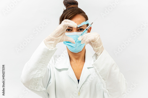 Médico com máscara azul e luvas cirúrgicas fazendo sinal de coração na altura dos olhos com a mão direita e a outra metade com a seringa da vacina. photo