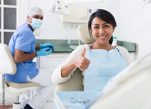 Happy latin american woman sitting in dental chair after teeth cure in modern clinic, gesturing thumbs up