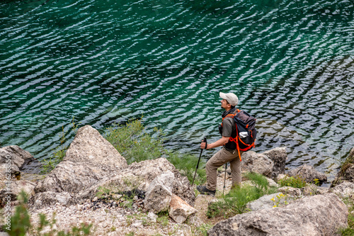 A tourist stands near the lake. A traveler with a backpack stands on the shore of the lake. A man in sportswear, a cap and with trekking poles. Climbing to the top of a mountain lake.