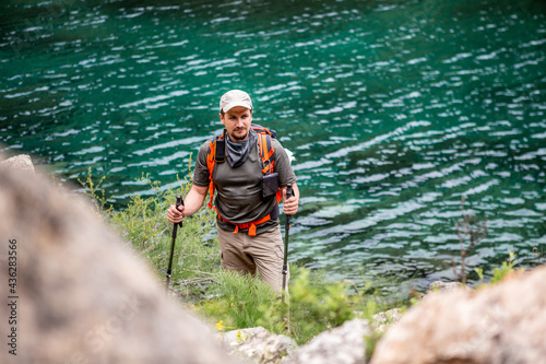 A tourist stands near the lake. A traveler with a backpack stands on the shore of the lake. A man in sportswear, a cap and with trekking poles. Climbing to the top of a mountain lake.