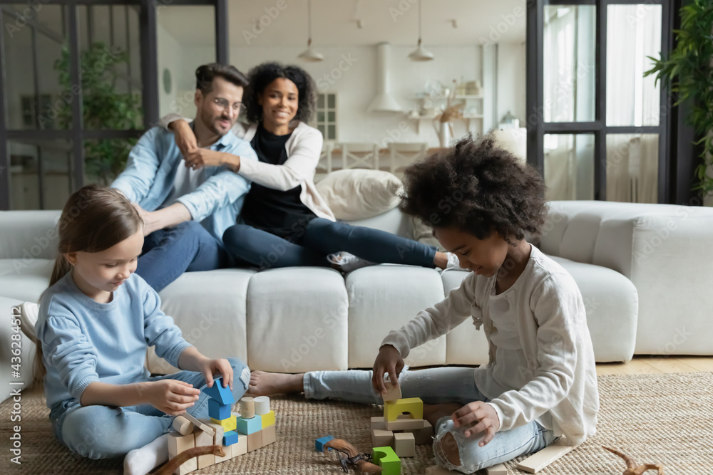 Happy young multiracial family with diverse small kids relax in cozy living room together. Smiling loving multiethnic mom and dad rest at home with little daughters. Adoption, rent concept.