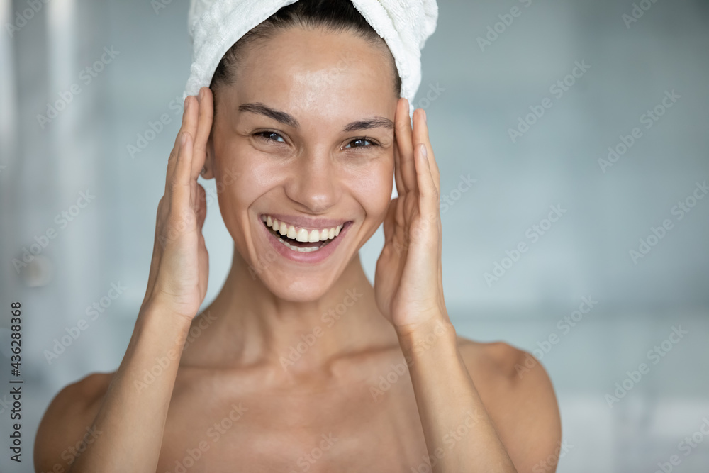 Head shot of happy woman with head wrapped in bath towel, touching face with toothy smile, applying cream, oil, lotion on facial skin after shower in bathroom. Skincare therapy concept. Portrait Head shot of happy woman with head wrapped in bath towel, touching face with toothy smile, applying cream, oil, lotion on facial skin after shower in bathroom. Skincare therapy concept. Portrait