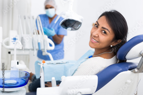 Cheerful hispanic female patient sitting in dental chair waiting for medical examination