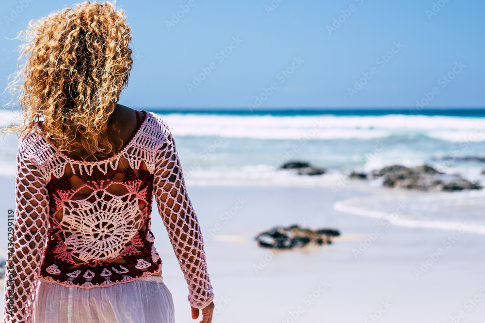 Back view of boho trendy female woman with lung curly blonde hair enjoying the beach alone looking at blue ocean waves in summer travel holiday vacation lifestyle activity