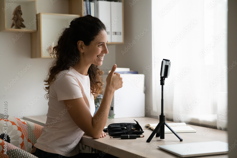 Happy beautiful millennial latin woman showing thumbs up gesture, recording advertising video in telephone, recommending makeup tools. Smiling hispanic female blogger enjoying vlogging activity. Happy beautiful millennial latin woman showing thumbs up gesture, recording advertising video in telephone, recommending makeup tools. Smiling hispanic female blogger enjoying vlogging activity.