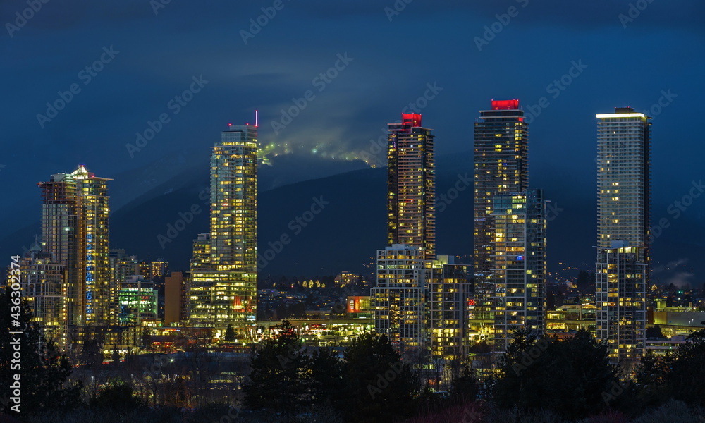 Fototapeta premium View of the new residential high-rise residential area in Burnaby City at night, against the backdrop of illuminated ski slopes on the mountain range