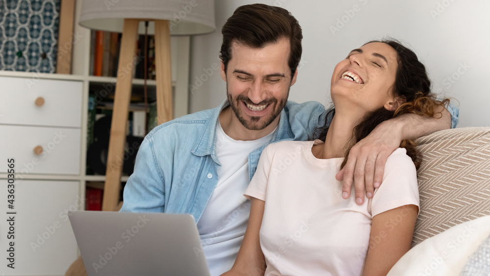 Joyful young married family couple looking at computer screen, watching funny video or comedian movie, laughing enjoying entertaining free leisure weekend time together, sitting on sofa at home. Joyful young married family couple looking at computer screen, watching funny video or comedian movie, laughing enjoying entertaining free leisure weekend time together, sitting on sofa at home.