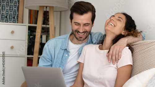 Joyful young married family couple looking at computer screen, watching funny video or comedian movie, laughing enjoying entertaining free leisure weekend time together, sitting on sofa at home.