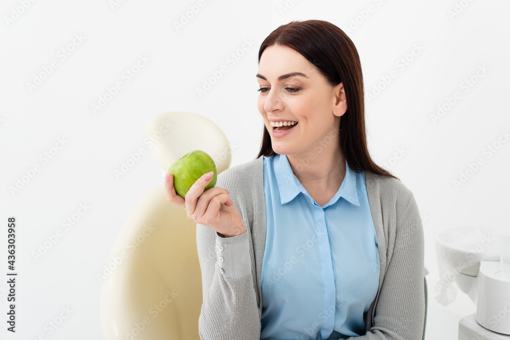smiling adult woman in dental chair looking at green apple in hand in clinic. smiling adult woman in dental chair looking at green apple in hand in clinic.
