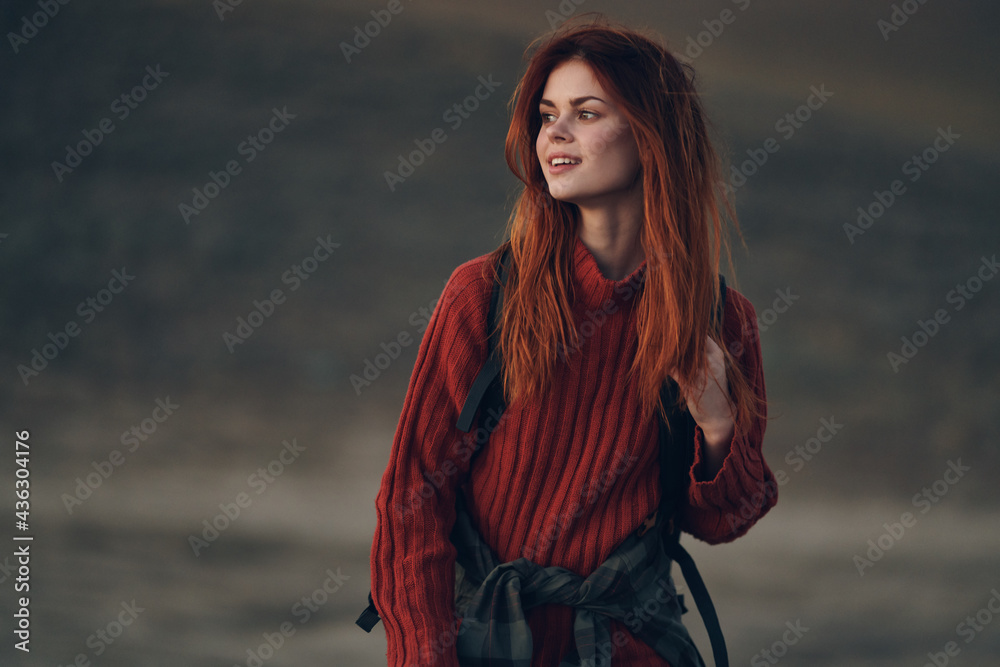 red-haired woman travels in nature in the mountains in a red sweater with a backpack on her back