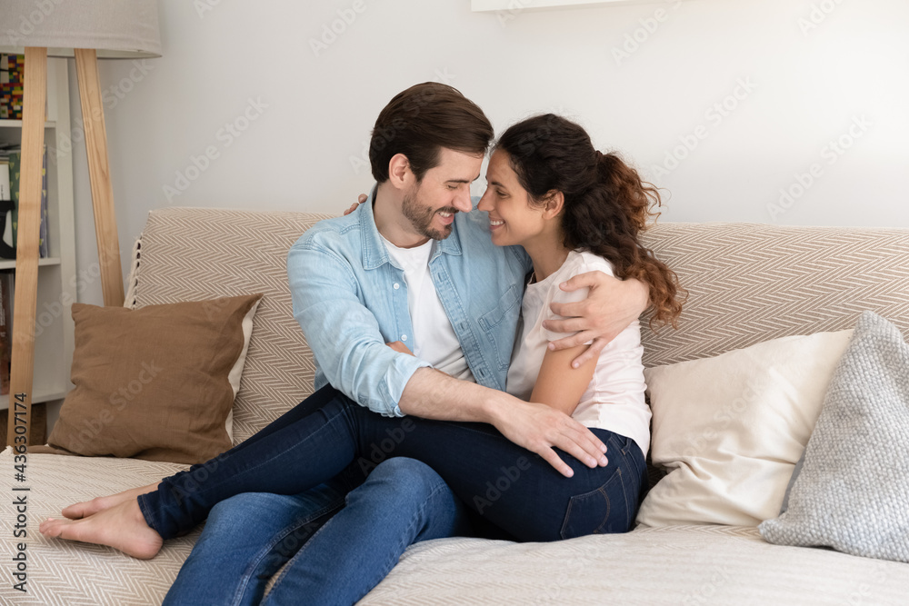 Happy affectionate bonding family couple cuddling sitting on comfortable sofa, showing sweet tender feelings at home. Smiling loving young man and woman enjoying lazy leisure weekend indoors.