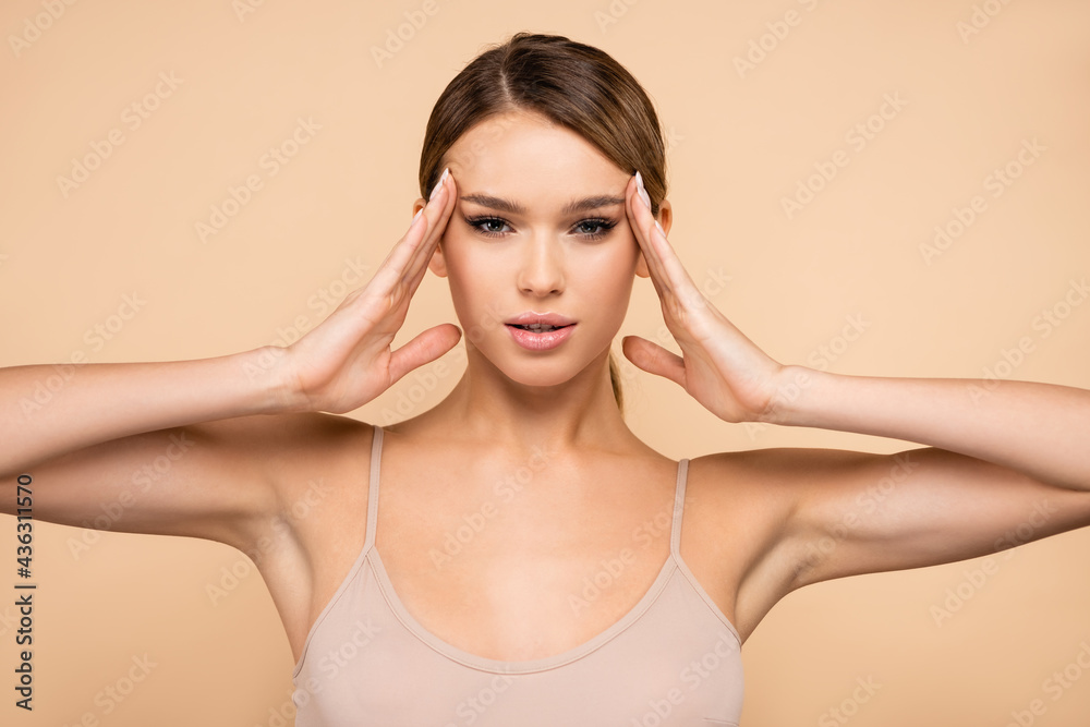 young woman looking at camera while holding hands near face isolated on beige.