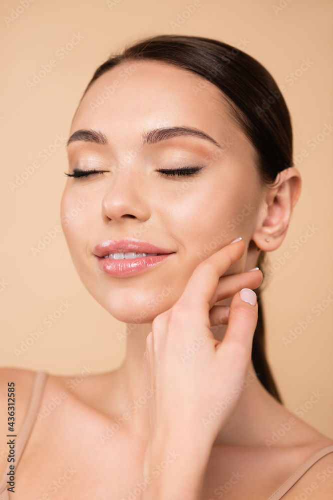 smiling woman with closed eyes touching face isolated on beige.