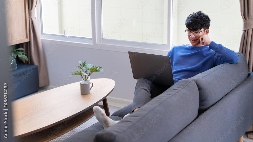 young attractive asian man relaxing comfortably working on laptop in bright living room at his home. young attractive asian man relaxing comfortably working on laptop in bright living room at his home.