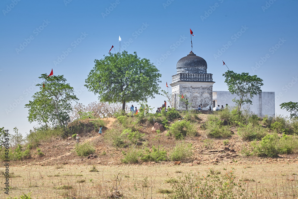 Small Hindu temple near Khajuraho. Temple stands on a hill. People and trees can be seen. Sky is blue. Day. Normal perspective.  