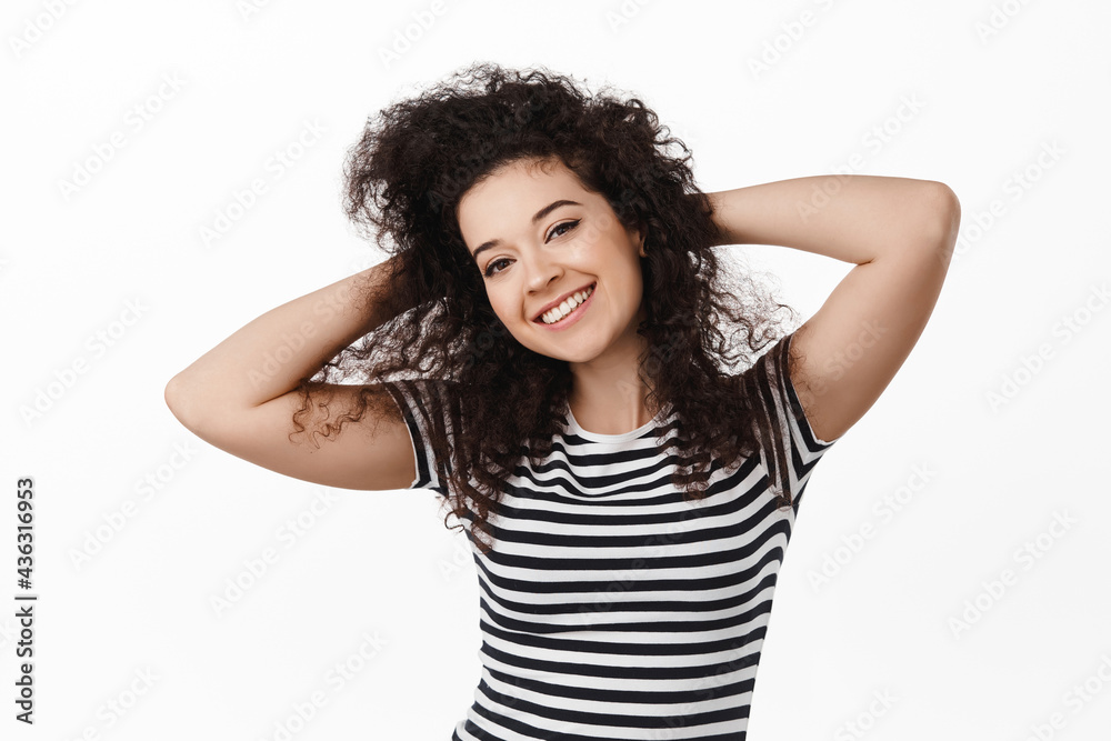 Portrait of happy brunette girl with curly natural hair, holding hands behind head, smiling and looking relaxed at camera, standing in t-shirt against white background Portrait of happy brunette girl with curly natural hair, holding hands behind head, smiling and looking relaxed at camera, standing in t-shirt against white background