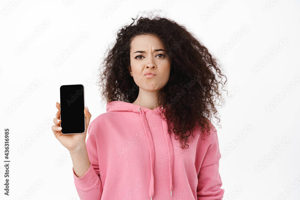 Skeptical brunette woman complaining, showing empty mobile screen and grimacing upset, disappointed by smartphone app, standing in pink hoodie against white background Skeptical brunette woman complaining, showing empty mobile screen and grimacing upset, disappointed by smartphone app, standing in pink hoodie against white background