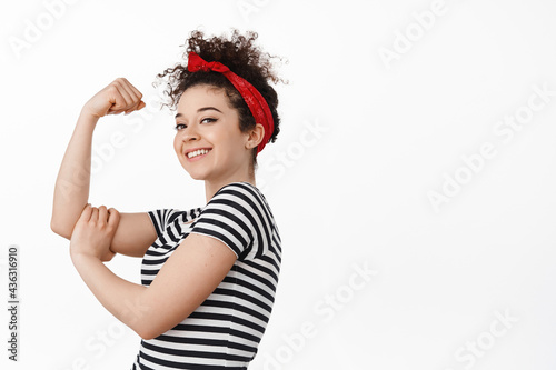 We can do it. Women power and feminism concept. Strong and confident brunette girl showing her arm muscle, flex biceps and smiling proud, standing against white background