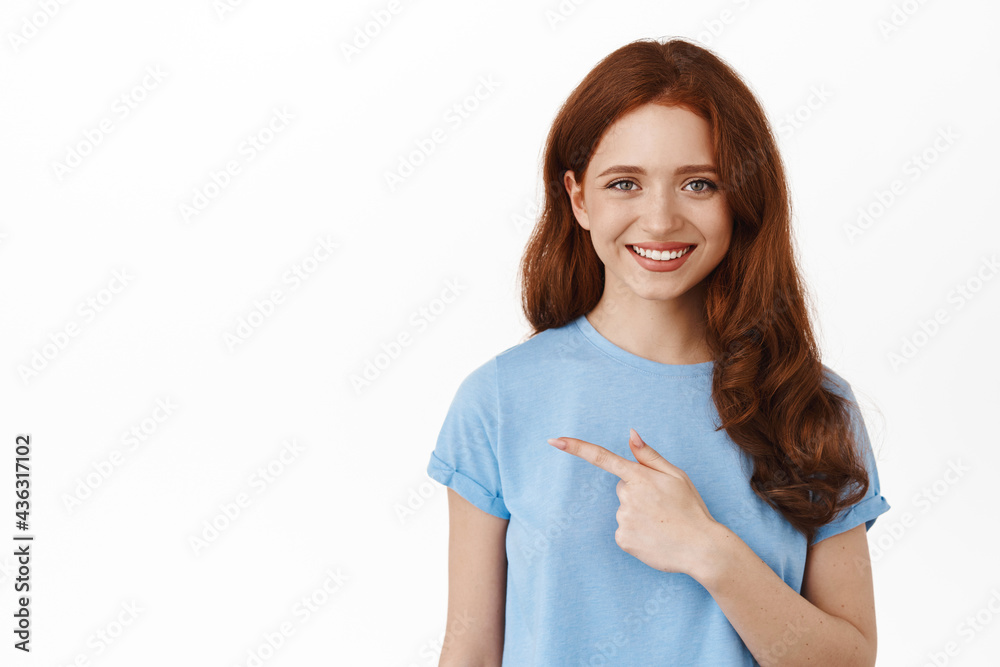 Young woman with red curly hair in t-shirt, pointing finger left, showing advertisement and smiling, recommending promotion, click link or logo, standing against white background Young woman with red curly hair in t-shirt, pointing finger left, showing advertisement and smiling, recommending promotion, click link or logo, standing against white background