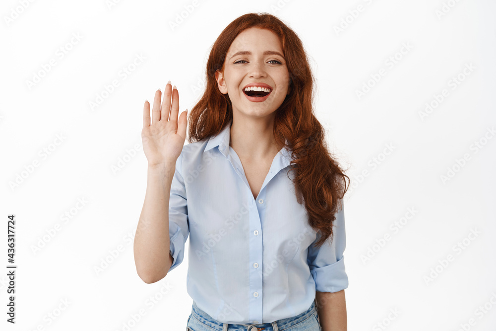Friendly redhead woman smiling happy and waving hand, say hello, make hi wave gesture, greeting a friend, standing in blouse against white background Friendly redhead woman smiling happy and waving hand, say hello, make hi wave gesture, greeting a friend, standing in blouse against white background