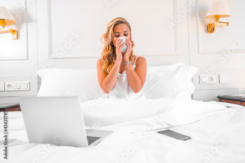 Portrait attractive young blond Caucasian woman in nightgown in hotel bed teleworking with computer. Working and traveling in a hotel