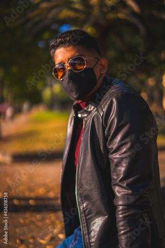 Fashion lifestyle, portrait of a young Latino in the city at a water fountain. Jeans, leather jacket and brown shoes. In a pandemic with a mask
