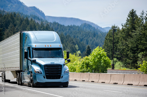 Blue bonnet big rig semi truck with black grille transporting cargo in refrigerator semi trailer running on the highway road with mountain and forest on background