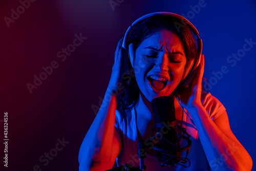 Caucasian woman in headphones singing into a microphone in neon light on a black background. An emotional girl is recording a song in a recording studio