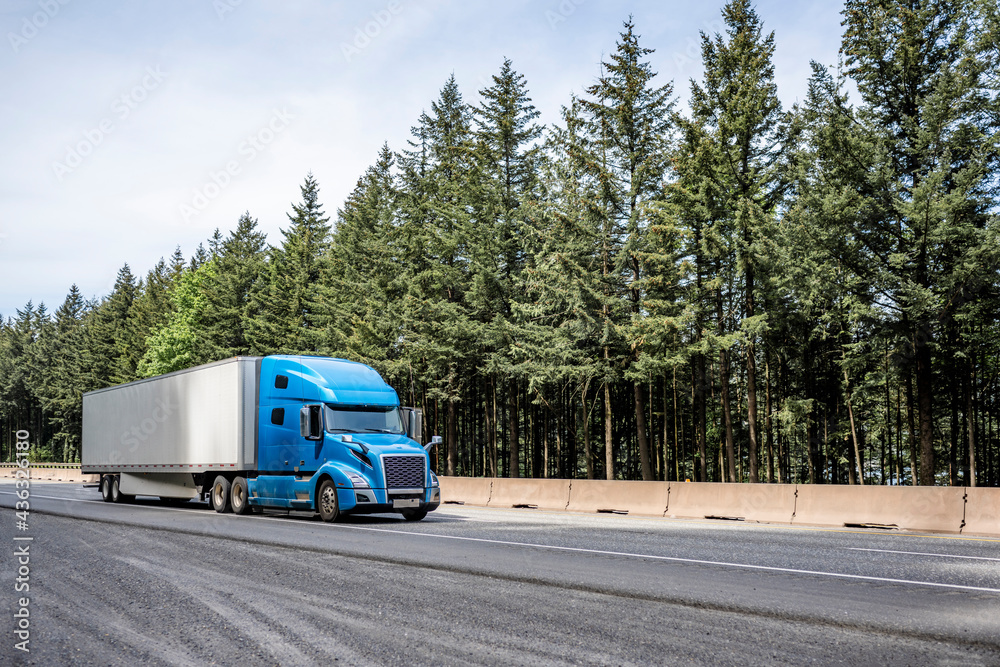 Stylish blue big rig long haul industrial semi truck transporting cargo in dry van semi trailer running on the highway road with pine trees on the side