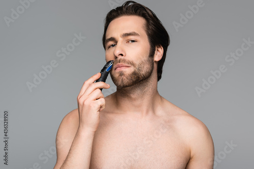 good-looking man trimming beard with electric trimmer isolated on grey.