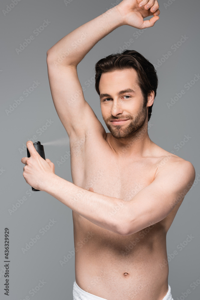 Fototapeta premium smiling and shirtless man with raised hand spraying antiperspirant isolated on grey.