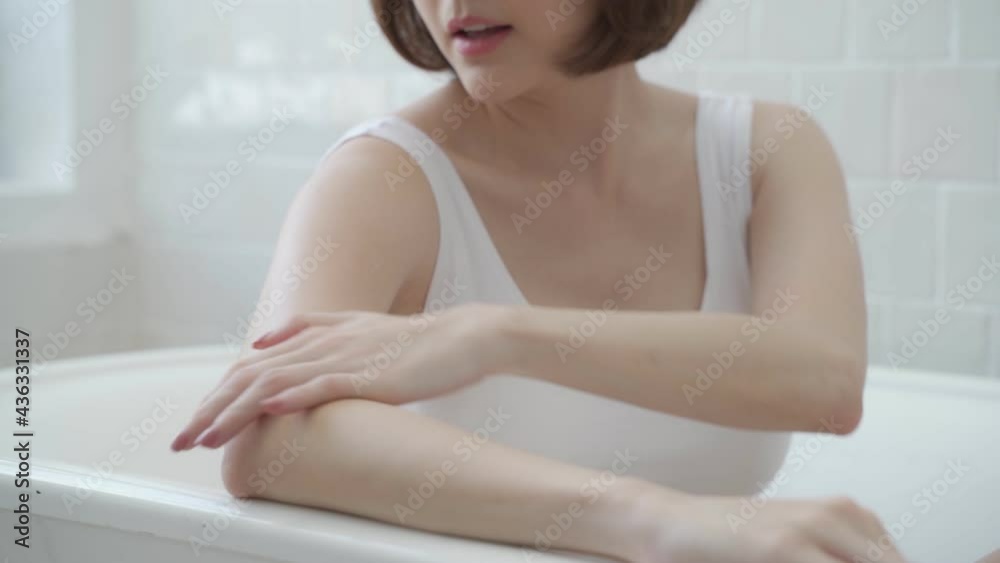 Asian woman short hair, applying cream lotion on hands, leg, and arm in white bathroom near bathtub at home studio decor some green plant. white cream rubbing to treatment skin condition. scrubbing Asian woman short hair, applying cream lotion on hands, leg, and arm in white bathroom near bathtub at home studio decor some green plant. white cream rubbing to treatment skin condition. scrubbing